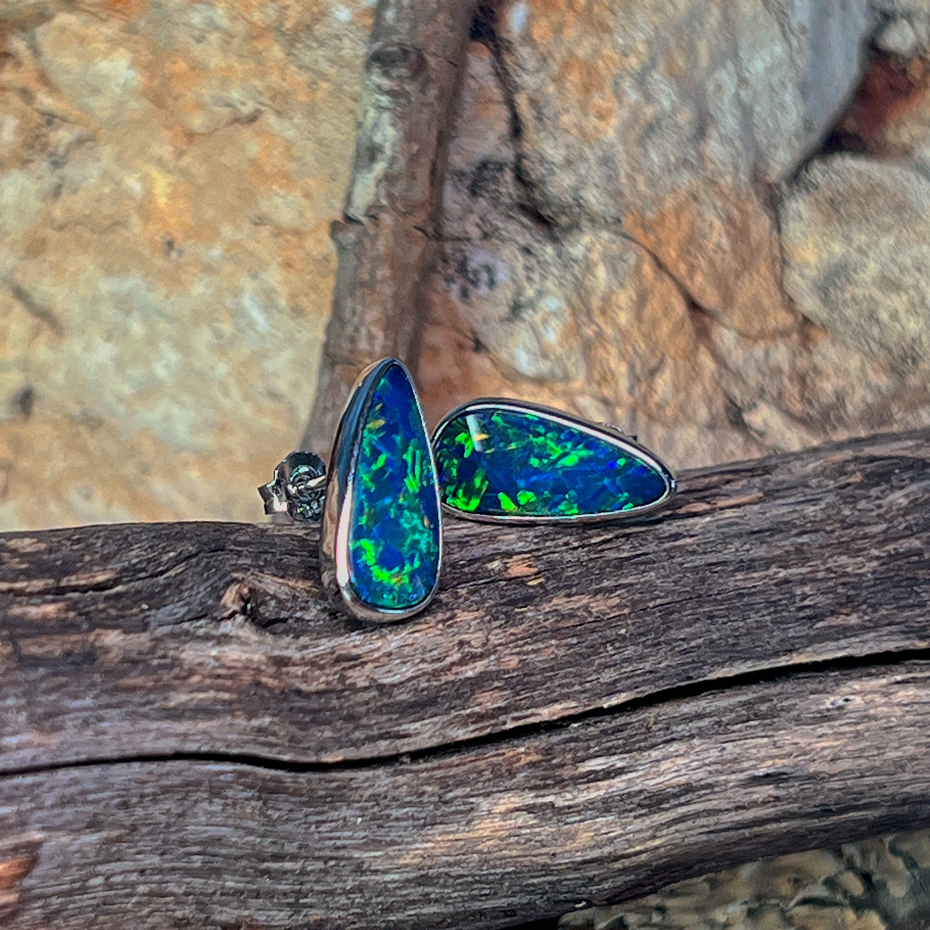 Silver bezel-set opal doublet stud earrings resting low on driftwood beside sandstone